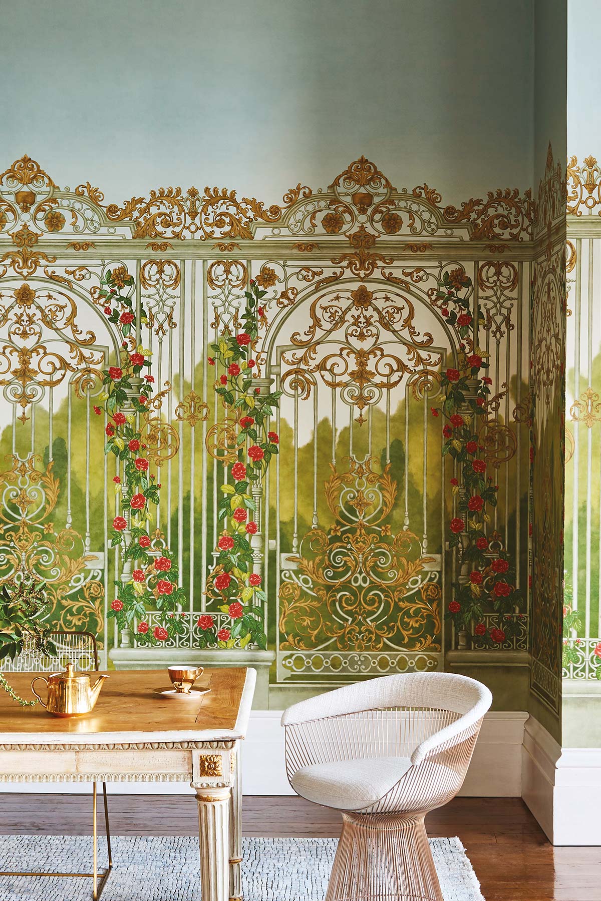 An ornate wall mural featuring gold filigree arches, green foliage, and red roses, shown in a styled interior with a white woven chair and a small table.
